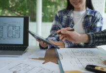 La tutela finanziaria è il primo pilastro di un progetto di successo Primo piano delle mani di due progettisti in studio, uno con camicia scozzese che usa una penna su un tablet digitale mostrato al collega, circondati da planimetrie e con un laptop che visualizza disegni architettonici. L'immagine rappresenta la fase cruciale della preventivazione e della selezione dei partner, ottimizzata con la verifica delle informazioni commerciali di Fatture in Cloud per la stabilità economica dello studio tecnico