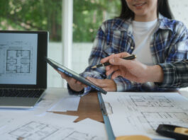 Primo piano delle mani di due progettisti in studio, uno con camicia scozzese che usa una penna su un tablet digitale mostrato al collega, circondati da planimetrie e con un laptop che visualizza disegni architettonici. L'immagine rappresenta la fase cruciale della preventivazione e della selezione dei partner, ottimizzata con la verifica delle informazioni commerciali di Fatture in Cloud per la stabilità economica dello studio tecnico