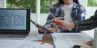Primo piano delle mani di due progettisti in studio, uno con camicia scozzese che usa una penna su un tablet digitale mostrato al collega, circondati da planimetrie e con un laptop che visualizza disegni architettonici. L'immagine rappresenta la fase cruciale della preventivazione e della selezione dei partner, ottimizzata con la verifica delle informazioni commerciali di Fatture in Cloud per la stabilità economica dello studio tecnico