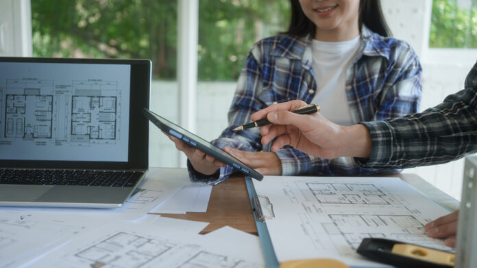 Architects reviewing house floor plans at desk, laptop and tablet in bright office, collaborating on home design project with blueprints and documents. Primo piano delle mani di due progettisti in studio, uno con camicia scozzese che usa una penna su un tablet digitale mostrato al collega, circondati da planimetrie e con un laptop che visualizza disegni architettonici. L'immagine rappresenta la fase cruciale della preventivazione e della selezione dei partner, ottimizzata con la verifica delle informazioni commerciali di Fatture in Cloud per la stabilità economica dello studio tecnico