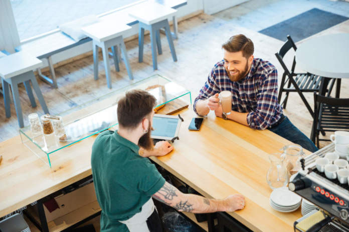 Cheeful man drinking coffee in cafeteria and talking to barista