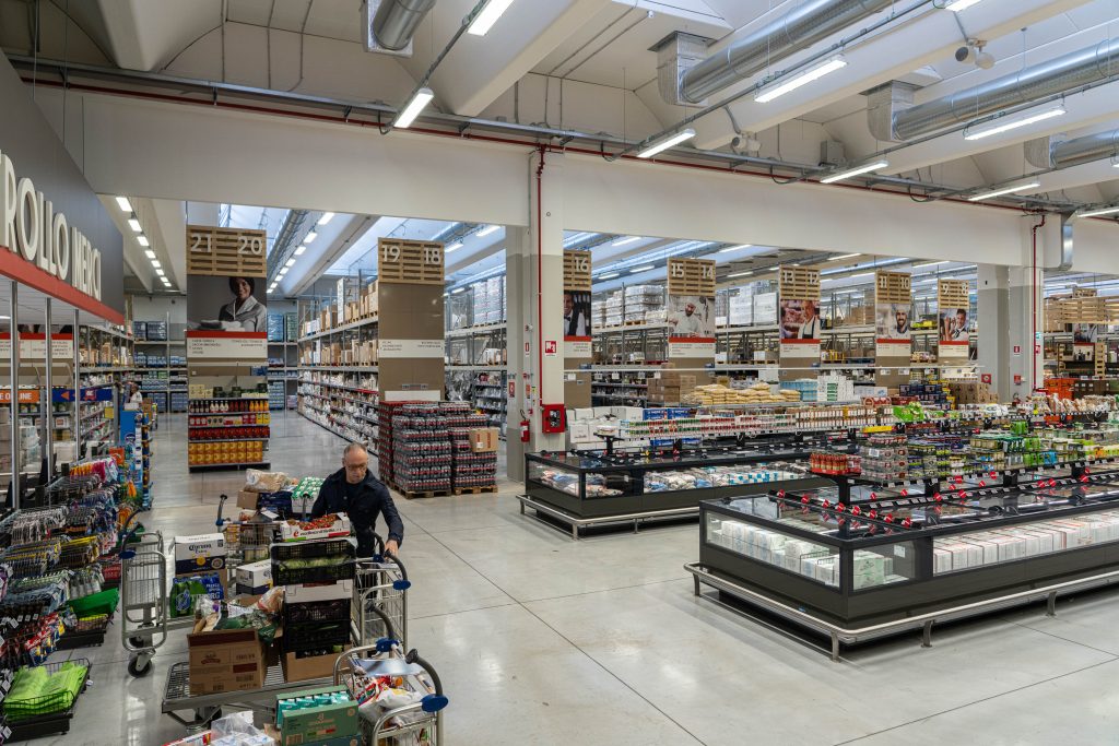 Interno di un cash & carry per bar con ampi scaffali, prodotti food & beverage e un operatore Horeca che rifornisce il carrello