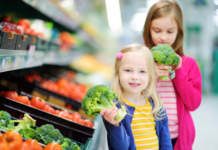 Quando lo stress dei genitori pesa sulla taglia dei figli Due bambine con in mano broccoli accanto al banco degli ortaggi di un supermercato