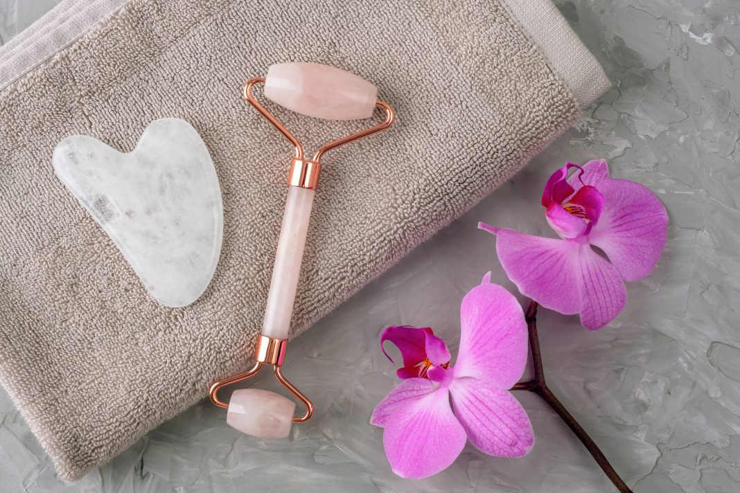 Rose quartz face roller and gua sha stone on towel with pink orchid on gray textured background. Selfcare concept. Top view, flat lay