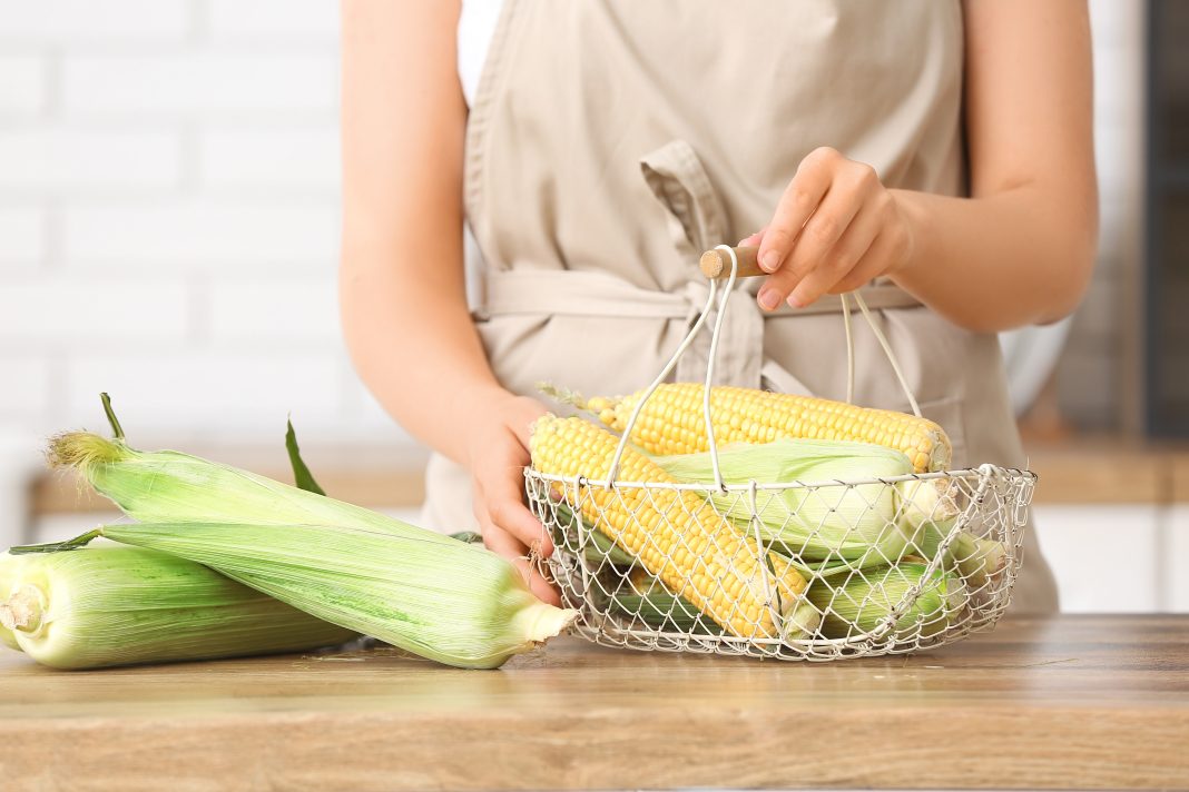 Woman holding basket with fresh corn cobs on wooden table in kitchen