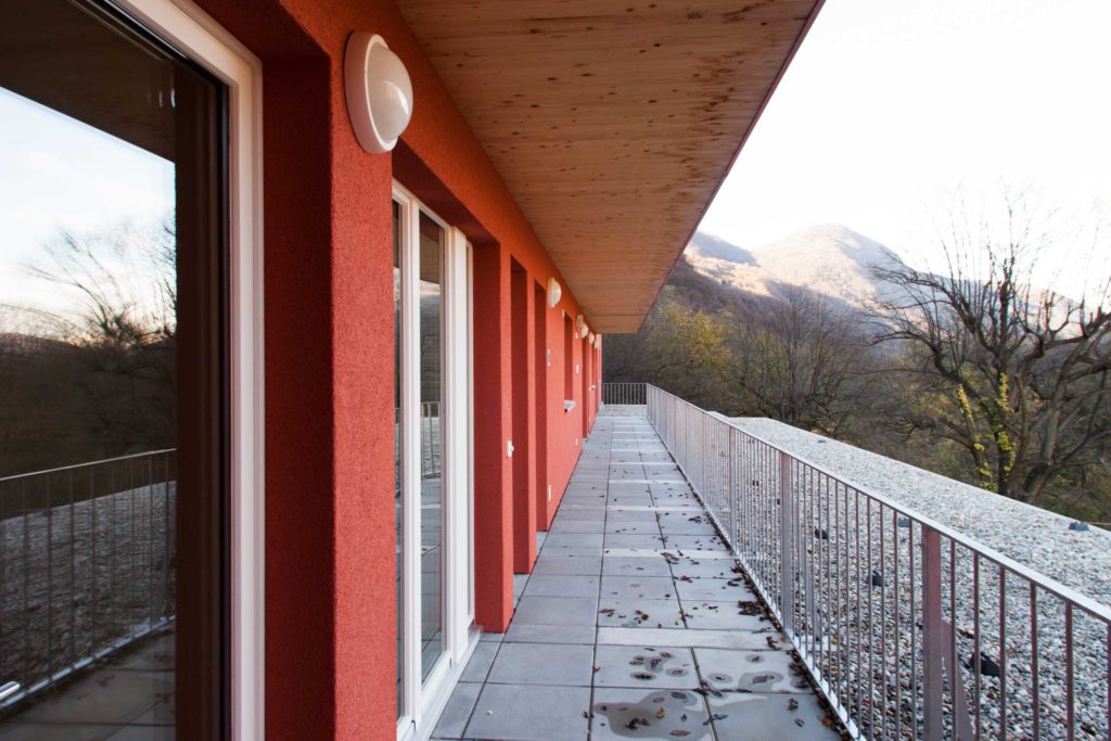Sbalzo in X-Lam e rivestimento a cappotto. Particolare del balcone che corre lungo tutta la facciata dell’edificio all’ultimo piano residenziale. Arretrato rispetto al filo delle facciate, ricorda gli storici ballatoi che nelle corti distribuivano gli accessi alle abitazioni. In questo caso l’affaccio diretto è sulla valle.