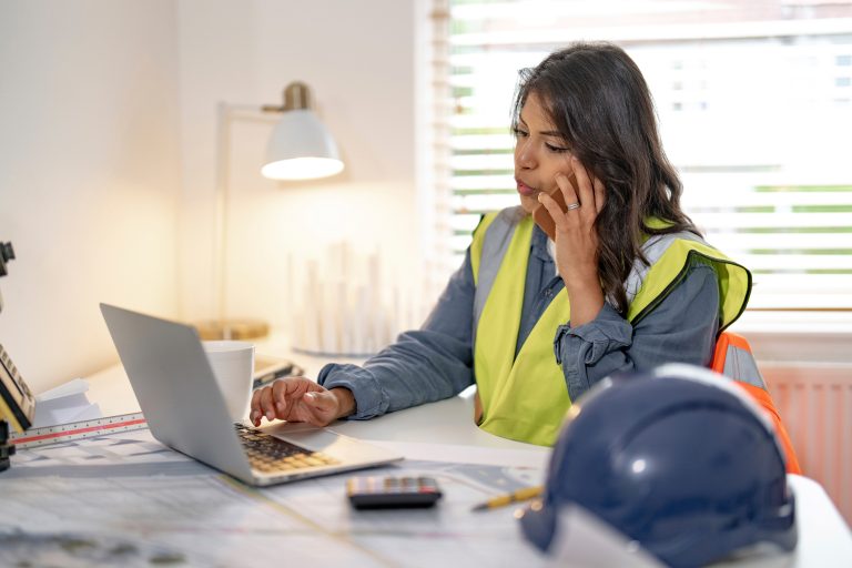 Donna con giubbotto ad alta visibilità al lavoro su laptop, mentre gestisce la fatturazione elettronica in una impresa edile.