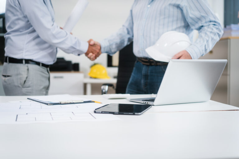 Scorcio di un ufficio tecnico con planimetrie, tablet, laptop e un casco antinfortunistico bianco in primo piano su una scrivania. Sullo sfondo, due professionisti in camicia si stringono la mano dopo un accordo, a simboleggiare la fiducia tra partner. L'immagine rappresenta l'importanza di basare le partnership e i contratti su solide informazioni commerciali in edilizia per prevenire rischi e insoluti nel settore delle costruzioni