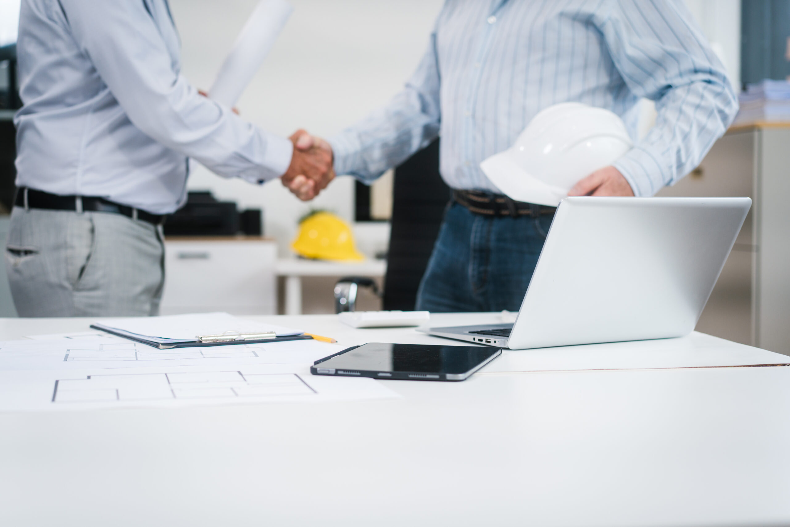 Scorcio di un ufficio tecnico con planimetrie, tablet, laptop e un casco antinfortunistico bianco in primo piano su una scrivania. Sullo sfondo, due professionisti in camicia si stringono la mano dopo un accordo, a simboleggiare la fiducia tra partner. L'immagine rappresenta l'importanza di basare le partnership e i contratti su solide informazioni commerciali in edilizia per prevenire rischi e insoluti nel settore delle costruzioni