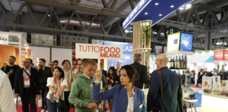 Interno dei padiglioni di TUTTOFOOD 2026 a Rho Fieramilano con visitatori tra gli stand e hostess che sorregge il cartello ufficiale della manifestazione