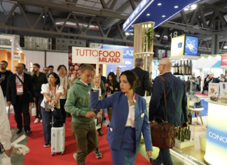Interno dei padiglioni di TUTTOFOOD 2026 a Rho Fieramilano con visitatori tra gli stand e hostess che sorregge il cartello ufficiale della manifestazione
