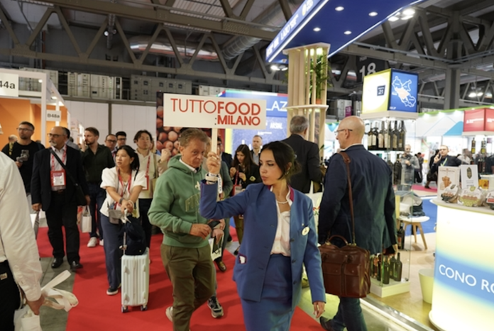 Tuttofood 2026 fiera di Rho Interno dei padiglioni di TUTTOFOOD 2026 a Rho Fieramilano con visitatori tra gli stand e hostess che sorregge il cartello ufficiale della manifestazione