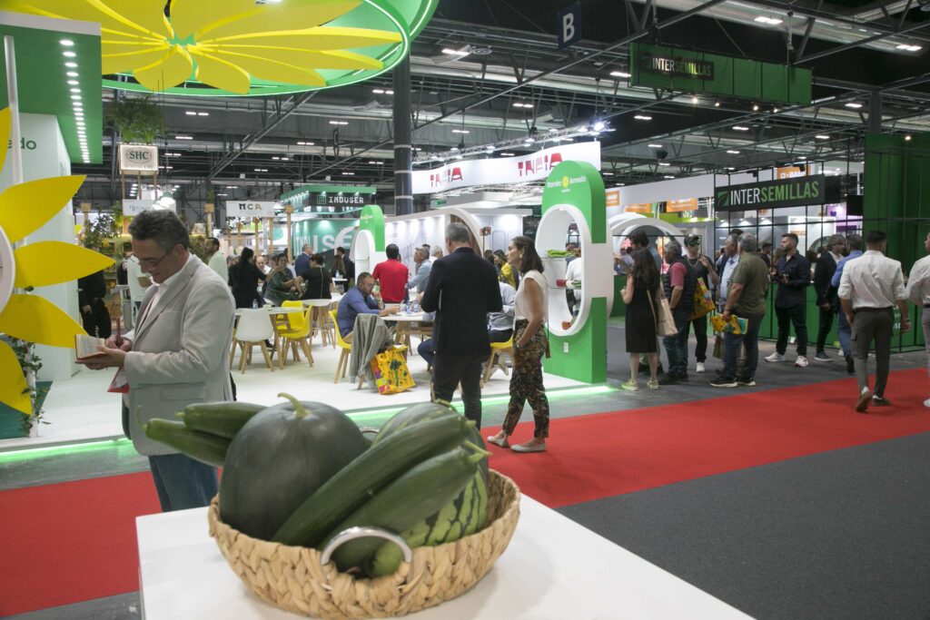 Dettaglio di un cesto di angurie e cetrioli in primo piano con lo sfondo del corridoio e degli stand espositivi aziendali alla fiera Fruit Attraction