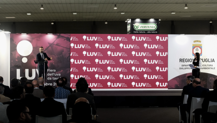 Un oratore parla dal podio durante una conferenza ufficiale a LUV Fiera dell'uva da tavola a Bari, con un grande fondale brandizzato e il logo della Regione Puglia sullo sfondo, davanti a una platea di operatori del settore