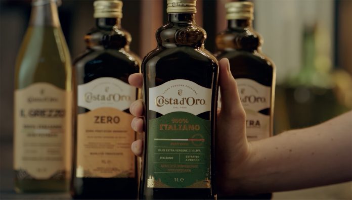 Bottiglia di olio extravergine di oliva Costa d’Oro 100% Italiano tenuta in mano, con altre referenze della linea sullo sfondo