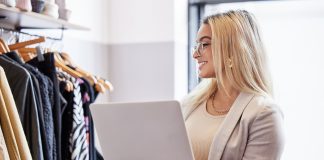 Addetta alla gestione magazzino in un negozio di abbigliamento controlla lo stock tramite laptop davanti a una fila di vestiti