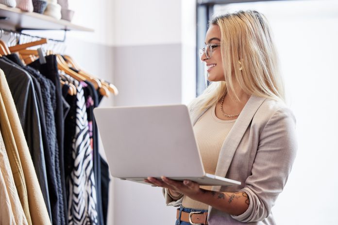 Addetta alla gestione magazzino in un negozio di abbigliamento controlla lo stock tramite laptop davanti a una fila di vestiti