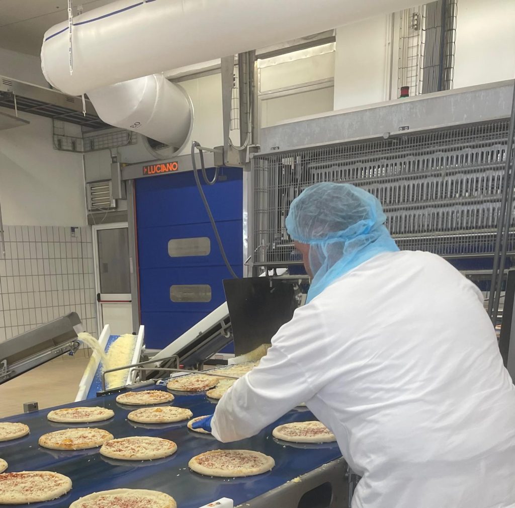 Operaio in camice bianco e cuffia blu al nastro trasportatore di pizze surgelate in uno stabilimento, con macchinari sullo sfondo impiegati nella gestione scarti di produzione