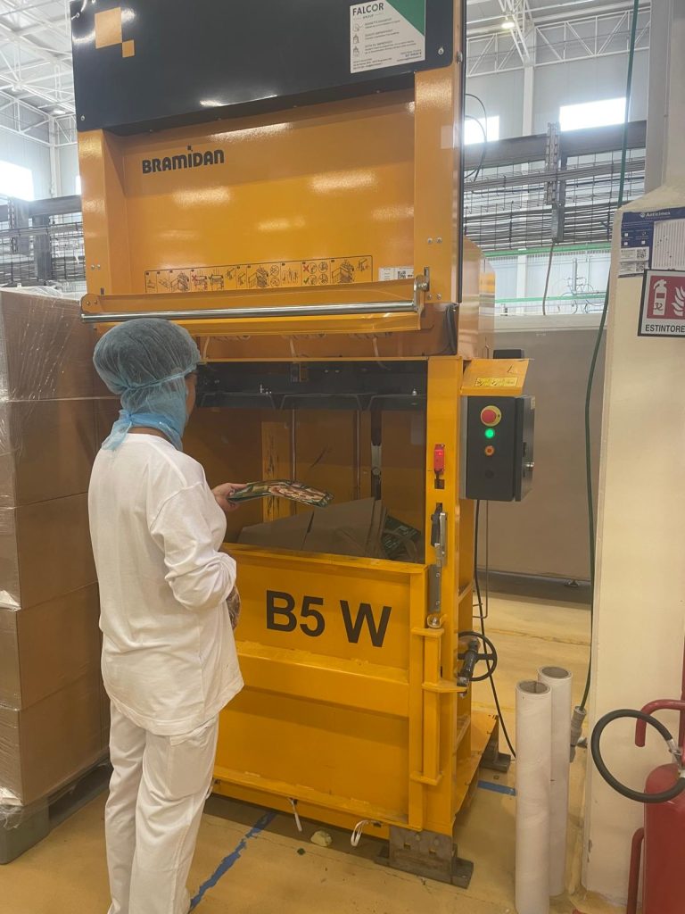Operaio in tuta bianca e cuffia blu che carica scarti di cartone in una pressa verticale gialla B5 W, nell’ambito della gestione scarti di produzione