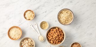 Mandorle gluten-free in diverse forme: ciotola centrale con mandorle intere, ciotole e cucchiaini con mandorle a scaglie, lamelle e farina di mandorle su un piano in marmo