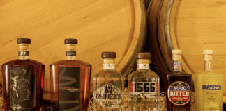 Ruffini Liquori: esposizione di sei bottiglie della gamma, inclusi gli amari Monumentale e Beatrix, i Gin 1566 e The Gin Brothers, Soul Bitter e Limoncello, con sfondo in legno e botte di rovere.