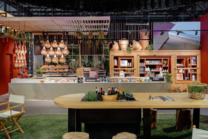 Tecnoarredamenti ospitalità: anco gastronomia e area degustazione in stile mercato contemporaneo, con prosciutti appesi, scaffali in legno e tavolo conviviale centrale che esprimono il nuovo format Tecnoarredamenti alla fiera HostMilano