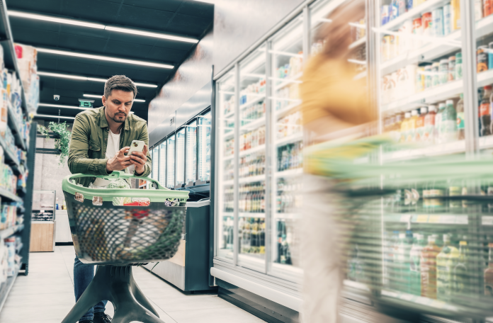iStock-2221746319 per Plastimark Uomo con smartphone fa la spesa utilizzando un carrello in plastica ergonomico, esempio delle soluzioni innovative e sostenibili che Plastimark sviluppa per la GDO moderna