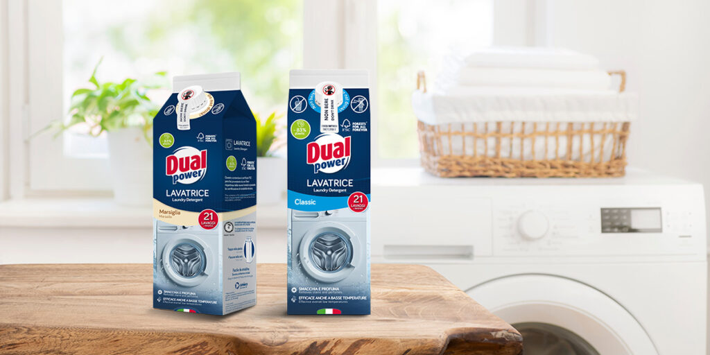 Detersivo Dual Power Lavatrice nelle varianti Classic e Marsiglia con nuovo packaging in carta riciclabile. I flaconi sono esposti in un ambiente lavanderia luminoso, con una lavatrice e un cesto di biancheria sullo sfondo, evidenziando il risparmio dell'83% di plastica