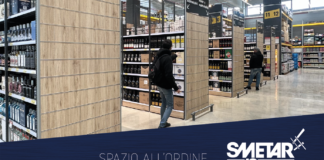 Un corridoio di supermercato allestito con scaffalature moderne Smetar con finiture in legno, piene di bottiglie di bevande alcoliche. Due persone osservano i prodotti, mostrando l’efficienza dell'organizzazione degli spazi espositivi per la GDO. In basso a destra è visibile il logo Smetar 40 Gruppo Metalsistem