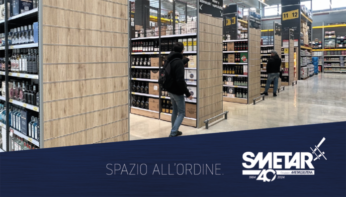 Smetar 2026 Un corridoio di supermercato allestito con scaffalature moderne Smetar con finiture in legno, piene di bottiglie di bevande alcoliche. Due persone osservano i prodotti, mostrando l’efficienza dell'organizzazione degli spazi espositivi per la GDO. In basso a destra è visibile il logo Smetar 40 Gruppo Metalsistem