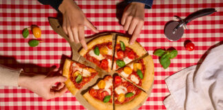 Dall'alto: mani che prendono fette di una Base Pizza Alimenta Produzioni con cornicione e pomodorini su tovaglia a scacchi rossa e bianca per Tuttofood 2026