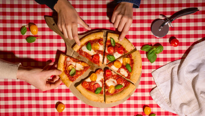 Dall'alto: mani che prendono fette di una Base Pizza Alimenta Produzioni con cornicione e pomodorini su tovaglia a scacchi rossa e bianca per Tuttofood 2026