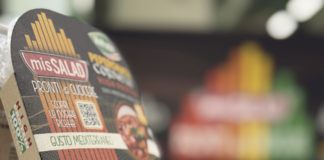 Alt Text: Dettaglio della confezione MisSalad "Peperonata Cornelio" High Protein di L’Insalata dell’Orto, linea High Protein 100% vegetale Pronti da Cuocere. Sullo sfondo sfocato, un'esposizione espositiva colorata a forma di piramide