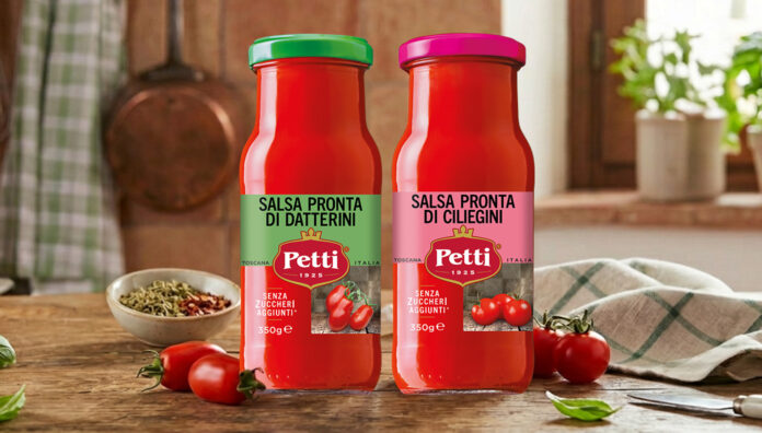 • Due bottiglie di vetro da 350g delle nuove Salse Pronte Petti, varianti Datterini e Ciliegini, presentate in anteprima da Petti a Tuttofood 2026. Le salse, con etichette chiare e tappi colorati (verde e fucsia), sono mostrate su un tavolo di legno in un ambiente di cucina rustica, circondate da pomodori freschi, basilico e una ciotola di spezie. L'immagine evidenzia la genuinità del prodotto 100% toscano