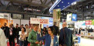 Una panoramica dell'affollata area espositiva di Tuttofood 2026 a Milano, con una donna in divisa blu che regge un cartello con la scritta "TUTTOFOOD: MILANO" tra i visitatori che percorrono un corridoio con tappeto rosso, stand fieristici e prodotti agroalimentari in mostra