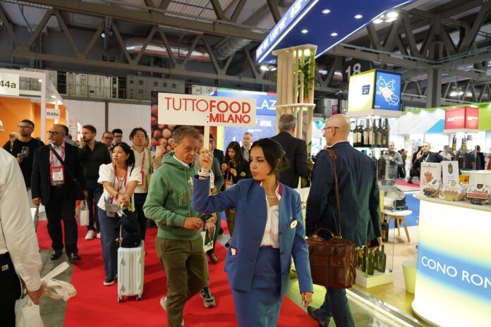 Una panoramica dell'affollata area espositiva di Tuttofood 2026 a Milano, con una donna in divisa blu che regge un cartello con la scritta 