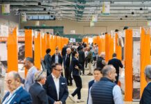 B2Cheese 2026: business e masterclass per la Gdo Panoramica all'interno del padiglione della Fiera di Bergamo dove si terrò l'evento B2B B2Cheese 2026, con stand espositivi arancioni e operatori della filiera casearia, inclusi buyer e professionisti della GDO, che interagiscono lungo il percorso espositivo della fiera verticale