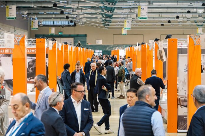 B2Cheese 2025 Panoramica all'interno del padiglione della Fiera di Bergamo dove si terrò l'evento B2B B2Cheese 2026, con stand espositivi arancioni e operatori della filiera casearia, inclusi buyer e professionisti della GDO, che interagiscono lungo il percorso espositivo della fiera verticale