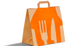 Alt Text: Una borsa della spesa in carta di colore marrone per il servizio Just Eat Grocery, posizionata contro uno sfondo nero. Sul lato anteriore della borsa è stampato un grande design geometrico di colore arancione. Al centro di questo design si trovano i profili bianchi stilizzati di una forchetta e di un coltello
