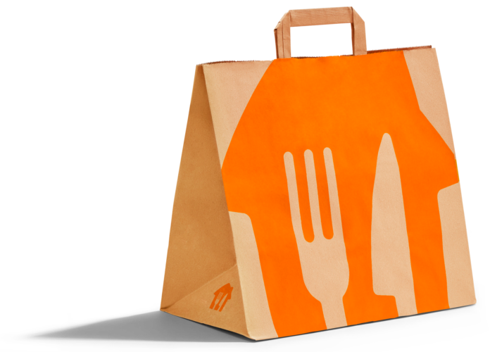 Alt Text: Una borsa della spesa in carta di colore marrone per il servizio Just Eat Grocery, posizionata contro uno sfondo nero. Sul lato anteriore della borsa è stampato un grande design geometrico di colore arancione. Al centro di questo design si trovano i profili bianchi stilizzati di una forchetta e di un coltello