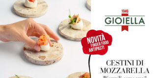 Mano che posiziona un cestino di mozzarella Gioiella farcito con gamberi e scorza di agrumi su alzata in pietra, circondato da altre monoporzioni gourmet e logo Gioiella con grafica Novità finger food antipasti