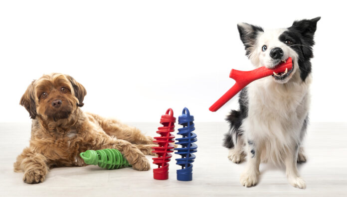 Due cani in posa su un pavimento chiaro con giochi colorati per animali domestici della linea Bamapet: a sinistra un cane marrone a pelo lungo è sdraiato accanto a un gioco verde, al centro ci sono due giochi a spirale verticali (uno rosso e uno blu), a destra un cane border collie bianco e nero tiene in bocca un gioco rosso a forma di ramo, su sfondo bianco