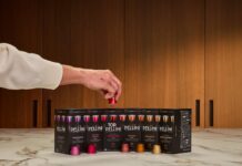 Una mano prende una capsula da una fila di nuove confezioni di capsule Pellini compatibili posizionate su un piano di marmo, con lo sfondo di una cucina moderna con pannelli in legno