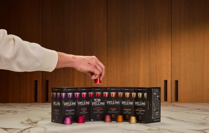 Capsule Pellini 2026 Una mano prende una capsula da una fila di nuove confezioni di capsule Pellini compatibili posizionate su un piano di marmo, con lo sfondo di una cucina moderna con pannelli in legno