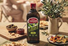 Pietro Coricelli: un rebranding che unisce contemporaneità e storia Bottiglia di Olio Extra Vergine di Oliva Classico Pietro Coricelli con la nuova grafica e packaging frutto del rebranding aziendale, posizionata su una tavola estiva con pietanze e rami di olivo