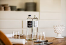San Benedetto Millennium Water: purezza millenaria, eleganza italiana Due bottiglie di acqua minerale San Benedetto Millennium Water in vetro, naturale e frizzante, posizionate su un tavolo apparecchiato in un ristorante di alto livello, con bicchieri e dettagli d'arredo sfocati sullo sfondo