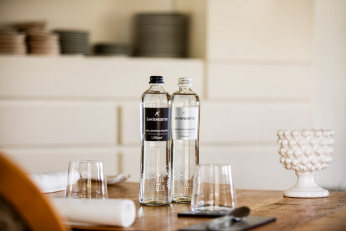 San Benedetto Millennium Water 1 2026 Due bottiglie di acqua minerale San Benedetto Millennium Water in vetro, naturale e frizzante, posizionate su un tavolo apparecchiato in un ristorante di alto livello, con bicchieri e dettagli d'arredo sfocati sullo sfondo