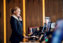 Come il gestionale aumenta la redditività degli hotel nel 2026 receptionist sorridente al telefono mentre utilizza un software gestionale per hotel alla reception di una struttura moderna e di design