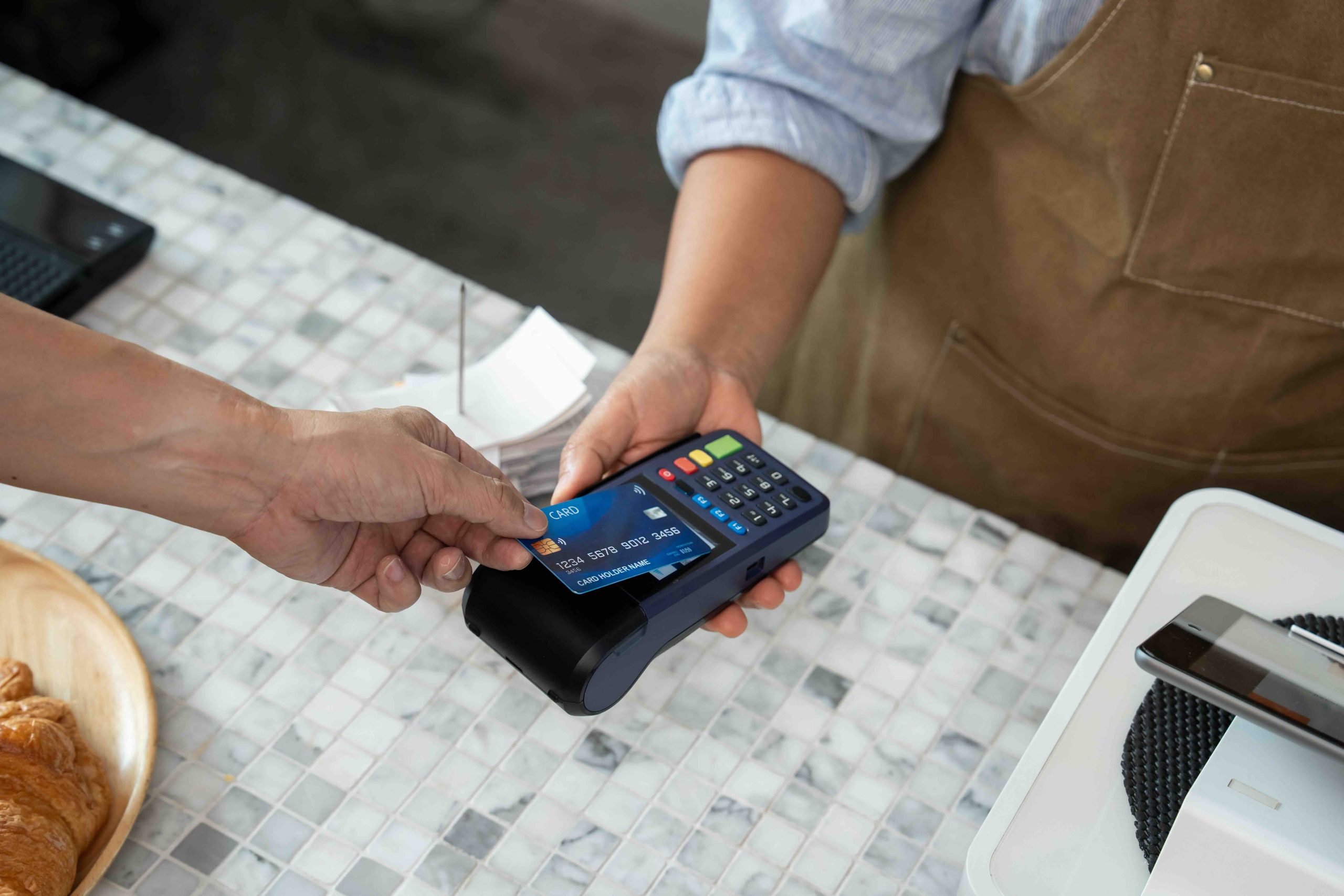 Customer hand tapping blue credit card with chip and wireless sign onto POS terminal held by barista wearing brown apron over marble table counter in cafe during contactless payment process