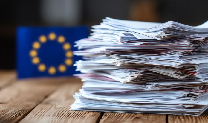 A large stack of disorganized papers sits on a wooden table with a blurred European Union flag in the background, symbolizing bureaucracy or administrative workload